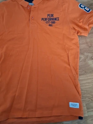Orange Peak Performance pikétröja - Snygg orange pikétröja från Peak Performance med tryck på bröstet och siffror på ärmen. Klassisk krage med två knappar och korta ärmar. Perfekt för en sportig och avslappnad stil.