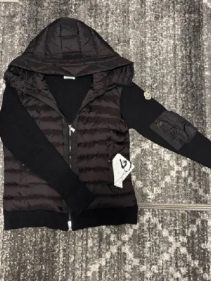 Svart cardiganjacka från Moncler S/M - Svart cardiganjacka från Moncler med quiltad front och huva, stickade ärmar och ribbade muddar. Dragkedja framtill och ficka med Moncler-logga på ärmen. Perfekt för lager-på-lager och snygg streetstil. Storlek S men funkar ändå lite med M