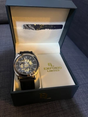 Emporio Limited svart klocka - Snygg klocka från Emporio Limited med svart armband och silverfärgat boett. Urtavlan har ett coolt skelettmönster där du ser de inre mekanismerna. Kommer i original ask med guldfärgad logga. Perfekt accessoar för dig som gillar stilrena detaljer.