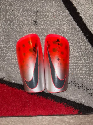 Nike benskydd röd och silver - Säljer ett par Nike benskydd i röd och silver med svart logga. De har en snygg gradientdesign och är lätta och smidiga att använda vid fotboll eller annan sport. Perfekta för dig som vill ha stilrena och funktionella benskydd.