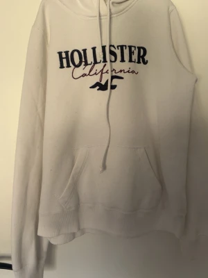 Vit hoodie från Hollister - Säljer en vit hoodie från Hollister med broderad text 'Hollister California' och logga på bröstet. Hoodien har huva med dragsko och en stor magficka framtill. Tillverkad i mjukt bomullsmaterial, perfekt för en avslappnad och stilren look.