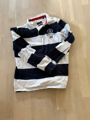 Randig rugbytröja från Henri Lloyd - Snygg rugbyinspirerad skjorta från Henri Lloyd med breda marinblå och vita ränder. Klassisk krage med röd insida, broderad logga och brittisk flagga på bröstet. Tillverkad i mjuk bomull och har långa ärmar med muddar.