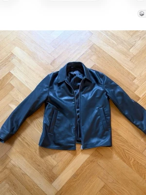 Svart klassisk skinnjacka - Stilren svart skinnjacka med rak passform och klassisk krage. Jackan har två snedställda fickor framtill och dragkedja. Perfekt för dig som gillar en tidlös och cool look.