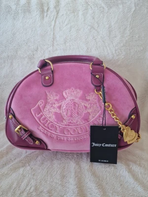 Rosa handväska från Juicy Couture - Superfin rosa handväska från Juicy Couture i mocka och skinnimitation med guldiga detaljer och kedja. Väskan har broderad logga framtill och hjärtberlock. Perfekt accessoar för att lyfta din outfit.