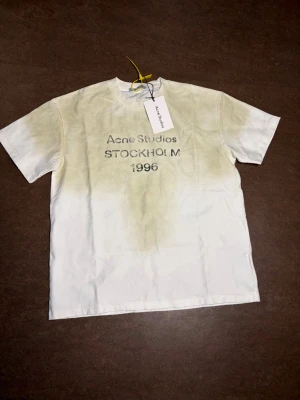 Vit t-shirt Acne Studios Stockholm - Vit t-shirt från Acne Studios med tryck 'Acne Studios STOCKHOLM 1996' på bröstet. Klassisk passform med rund hals och korta ärmar. Perfekt för dig som gillar stilrena och trendiga plagg med en touch av streetwear.