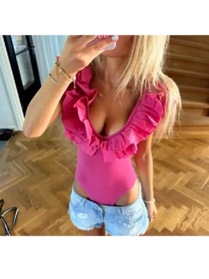 Rosa body med volangdetaljer - Snygg rosa body med djup v-ringning och stora volanger längs axlar och urringning. Tight passform och högt skurna ben. Perfekt för dig som vill sticka ut med en trendig och feminin look.