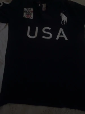 Mörkblå Polo Ralph Lauren skjorta - Säljer en mörkblå skjorta från Polo Ralph Lauren med stora vita bokstäver 'USA' på bröstet och ryggen, samt nummer 10 och amerikansk flagga på ryggen. Klassisk polokrage och knappdetalj framtill. Ikoniska Polo-loggan broderad på bröstet.