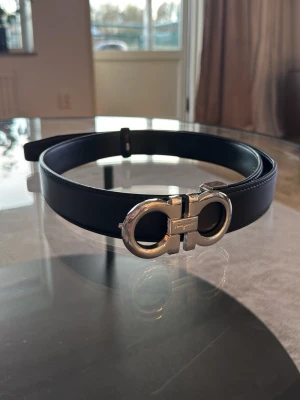 Svart skärp från Salvatore Ferragamo 115cm - Stilrent svart skärp från Salvatore Ferragamo med en ikonisk, rundad silverspänne. Skärpet är gjort i slätt skinn och har en elegant design som lyfter vilken outfit som helst. 115cm