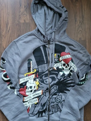Grå Ed Hardy hoodie med tryck - Säljer en grå zip-hoodie från Ed Hardy med stora, färgglada tryck av dödskallar, hjärtan och texter som 'Love Kills Slowly' och 'Live Fast'. Hoodien har huva med dragsko och tryck även på ärmarna. Perfekt för dig som gillar streetwear och statement-plagg.