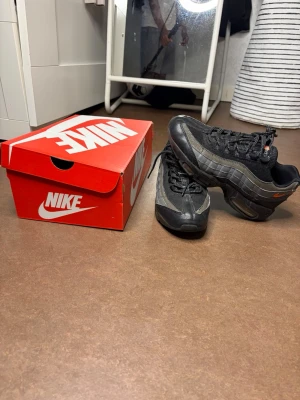 Nike Air Max 95 svarta sneakers - Nike Air Max 95 sneakers i svart och grått med orange detaljer. Klassisk vågig design, synlig Air-dämpning i sulan och robust mesh- och läderöverdel. Snygga snörningar och ikoniska loggor på häl och tunga. Perfekta för dig som gillar streetwear.