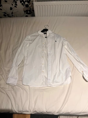 Vit skjorta från Polo Ralph Lauren - Klassisk vit skjorta från Polo Ralph Lauren med button-down krage och broderad logga på bröstet. Skjortan har slim fit passform, långa ärmar och knäppning framtill. Perfekt för en stilren och tidlös look.