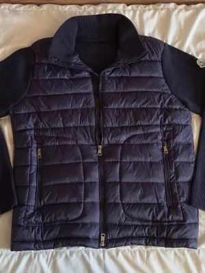 Moncler cardigan  - Snygg mörkblå pufferjacka från Moncler med stickade ärmar och krage. Jackan har två dragkedjeförsedda fickor framtill och quiltad front. Perfekt för kalla dagar och ger en sportig men stilren look.