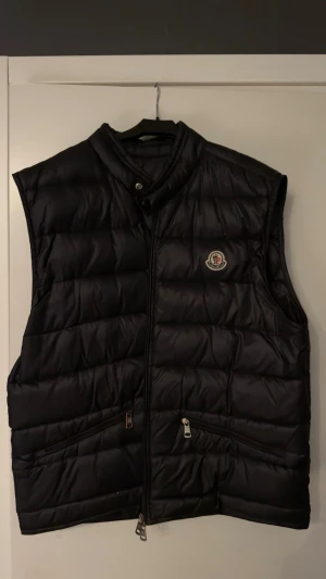 Moncler gui väst - Snygg svart dunväst från Moncler med hög krage och dragkedja framtill. Västen har två snedställda fickor med dragkedja och Moncler-logga på bröstet. Perfekt för lager-på-lager och håller dig varm med sin fluffiga dunfyllning.