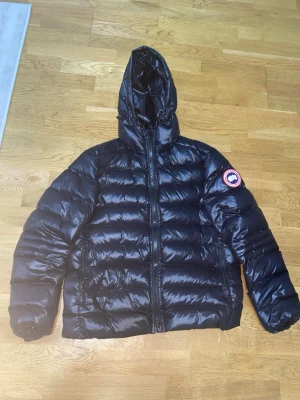 Svart dunjacka från Canada Goose - Snygg svart dunjacka från Canada Goose med glansig finish, huva och dragkedja framtill. Jackan har märkets klassiska logga på ärmen och invändiga axelband med Canada Goose-tryck. Perfekt för kalla dagar.