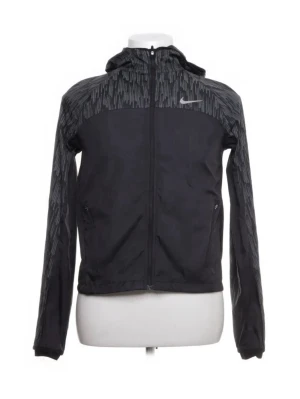 Svart Nike vindjacka med huva - Svart vindjacka från Nike med hel dragkedja, huva och diskret mönster i grått över axlar och ärmar. Jackan har två fickor med dragkedja och en liten Nike-logga på bröstet. Perfekt för blåsiga dagar och sportiga outfits. Skriv för mer bilder 