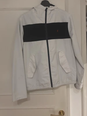 Vit och svart vindjacka från Polo Ralph Lauren - Snygg vit vindjacka med svart bröstpanel och röd logotyp från Polo Ralph Lauren. Jackan har huva, dragkedja framtill och två snedställda fickor. Perfekt för att hålla vinden borta och ge en clean look.