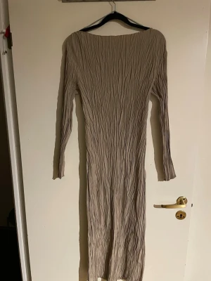 Beige långklänning med struktur - Säljer en beige långklänning med snygg strukturerad yta och långärmad design. Klänningen har en kroppsnära passform och är tillverkad i ett syntetmaterial som ger ett coolt, veckat utseende. Perfekt för dig som gillar stilrena och moderna plagg.