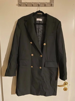 Svart lång kavaj med guldknappar - Stilren svart kavaj från MNG med dubbelknäppning och guldiga knappar. Kavajen har klassisk krage, två framfickor och är längre i modellen. Materialet är slätt och fodrat för extra komfort. Perfekt för dig som gillar en elegant och modern look.