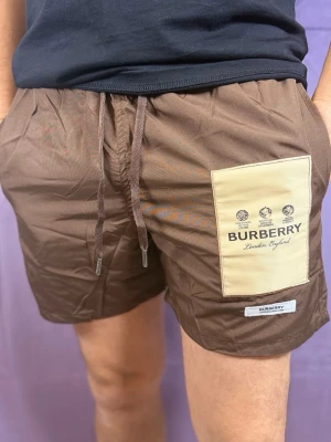 Bruna Burberry shorts med patch - Bruna shorts från Burberry med beige patch på höger ben där Burberry-loggan och texten 'London England' är tryckt. Shorts med dragsko i midjan och avslappnad passform. Materialet ser ut att vara lätt och bekvämt, perfekt för sommaren.