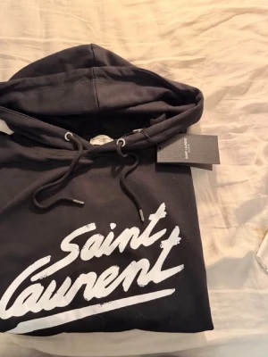 Svart Saint Laurent hoodie med tryck - Svart hoodie från Saint Laurent med stor vit logotyp framtill i graffiti-stil. Klassisk huva med dragsko och ribbade muddar. Mjuk bomullskänsla och streetwear-vibe, perfekt för dig som gillar exklusiva märken och snygg design.