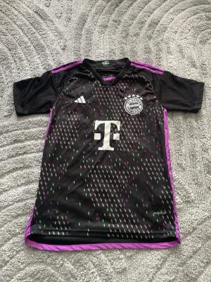 FC Bayern München Adidas matchtröja - Säljer en svart FC Bayern München matchtröja från Adidas i storlek S. Tröjan har lila och gröna detaljer, vita loggor och klubbmärke på bröstet samt lila ränder på axlarna. Slim fit-modell i funktionsmaterial, perfekt för träning eller match.