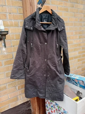 Svart parkas med bälte från Herluf Design stl 38 - Lång svart parkas från Lovely med avtagbart bälte i midjan, dragkedjor och tryckknappar framtill samt flera fickor med dragkedja. Jackan har hög krage och justerbar huva, perfekt för kyliga dagar. Snygg och funktionell design.