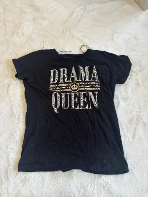 Svart t-shirt Drama Queen 146-153 - Svart t-shirt från Drama Queen med stort vitt och zebramönstrat tryck på bröstet och gulddetaljer. Klassisk rund hals och korta ärmar. Perfekt för dig som vill sticka ut med en cool statement-tee.