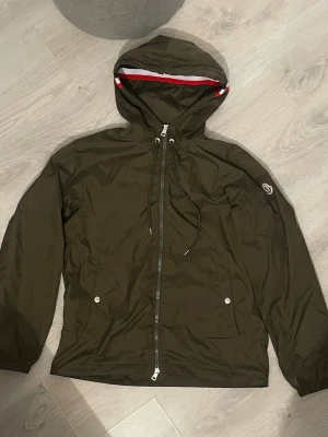 Moncler windbreaker  - Snygg olivgrön vindjacka från Moncler med huva och dragsko. Jackan har hel dragkedja framtill, två fickor med tryckknappar och Moncler-logga på ärmen. Insidan av huvan har en röd, vit och blå rand. Perfekt för blåsiga dagar.