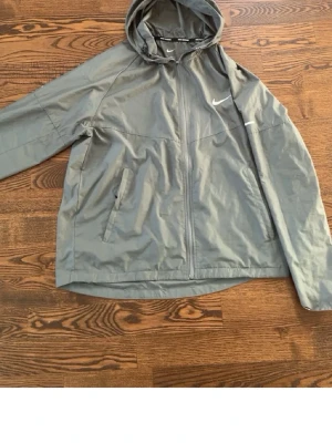  Nike Miller - Snygg grå vindjacka från Nike med dragkedja framtill och två fickor med dragkedja. Jackan har en praktisk huva och den klassiska Nike-loggan på bröstet. Perfekt för blåsiga dagar och enkel att matcha med olika outfits.