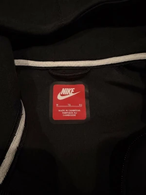 Svart Nike hoodie med dragkedja - Snygg svart hoodie från Nike med dragkedja framtill och klassisk logga på bröstet. Hoodien har huva och är tillverkad i mjukt material som passar perfekt till en avslappnad streetwear-look.