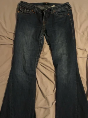Blå bootcut jeans från True Religion - Säljer ett par blå bootcut jeans från True Religion med klassiska kontrastsömmar och snygga fickdetaljer bak. Jeansen har låg midja och är tillverkade i denim med lätt tvättad look. Perfekta för dig som gillar en avslappnad men trendig stil. Lite defekter nertill men inget som syns när man har på sig de💓