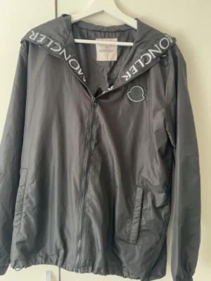 Grå/svart vindjacka  - Snygg grå vindjacka från med huva och dragkedja. tryck på huvan. Två fickor framtill och lätt material som passar perfekt till blåsiga dagar.