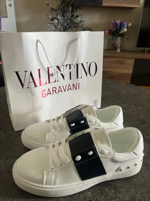 Valentino Garavani Skor - Snygga vita sneakers från Valentino Garavani med bred svart rem över vristen och silverfärgade nitar på hälen. Skorna har vita snören, platt sula och är tillverkade i slätt läder. Perfekta för dig som vill ha en stilren och exklusiv look.