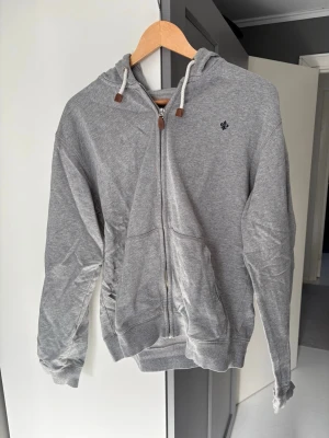 Grå zip-hoodie med broderad logga - Snygg grå hoodie med dragkedja och huva med vita snören och bruna detaljer. Broderad liten logga på bröstet och två stora fickor framtill. Tillverkad i mjukt bomullsmaterial, perfekt för en avslappnad stil.