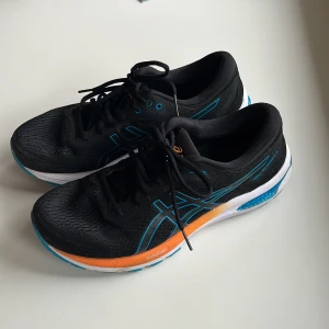Svarta löparskor från Asics - Säljer ett par svarta Asics löparskor med blå och orange detaljer. Skorna har snörning och en vit sula med blått mönster undertill. Perfekta för löpning och träning. De är sparsamt använda! 🤗
