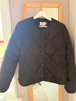 Svart quiltad pufferjacka från Nelly  - Stilren svart pufferjacka från nelly med quiltat mönster och rund halsringning. Jackan har tryckknappar framtill och två sidofickor. Perfekt för kalla dagar när du vill hålla värmen och se snygg ut.