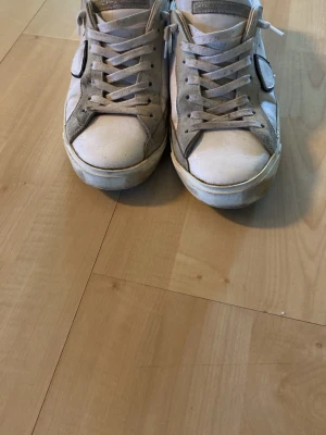 Vita Philippe Model sneakers med svart detalj - Snygga vita sneakers från Philippe Model med svart häl och ikonisk sköld på sidan. Skorna har klassisk låg profil, vita snören och detaljer i mocka och läder. Perfekta för en stilren och trendig look.