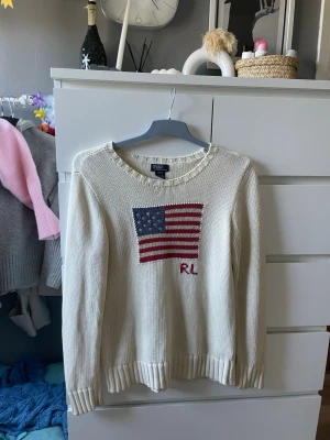 Vit stickad tröja Polo Ralph Lauren - Vit stickad tröja från Polo Ralph Lauren med amerikansk flagga och RL-broderi på bröstet. Tröjan har rund halsringning, ribbade muddar och är gjord i 100% bomull. Perfekt för en klassisk och avslappnad stil. I barn storlek är det L vilket passar 14-16 ish/ S-M. Bara av att höra av er om något🥰