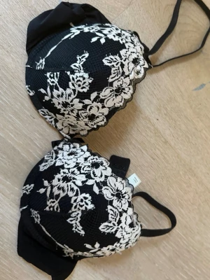 Svart spets-bh med blommönster Lindex - Snygg svart bh från Lindex med vit spets och blommigt mönster. Bh:n har vadderade kupor och justerbara axelband. Märkt med modellen MALVA. Perfekt för dig som gillar feminina detaljer och klassisk design.