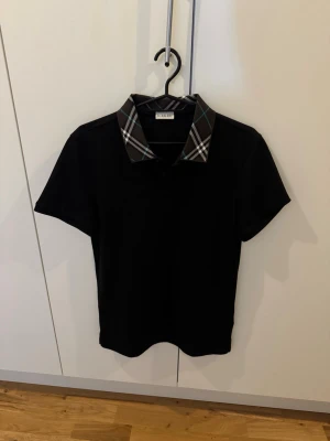 Svart Burberry topp med rutig krage - Snygg svart topp från Burberry med klassisk rutig krage i brunt, vitt och blått. Kortärmad modell med normal passform, perfekt för en stilren och trendig look. Ikonisk detalj med Burberrys signaturrutor på kragen.