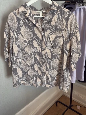 Kortärmad skjorta med ormmönster - Snygg kortärmad skjorta från H&M med grått och beige ormmönster. Skjortan har klassisk krage, bröstficka och avslappnad passform. Perfekt för dig som gillar trendiga mönster och vill sticka ut lite extra.