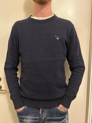 Marinblå stickad tröja från GANT - Säljer en klassisk marinblå stickad tröja från GANT med rund halsringning och broderad logga på bröstet. Tröjan har långa ärmar och ribbade muddar vid ärmslut och nederkant.