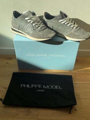 Grå Philippe Model sneakers i mocka - Snygga grå sneakers från Philippe Model i mocka och läder med vita detaljer och vita skosnören. Klassisk låg modell med diskret logga på sidan och bekväm sula. Perfekta för en stilren och trendig look.