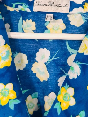 Blommig blå klänning Laura Bertucchi - Färgstark klänning från Laura Bertucchi med blå botten och blommigt mönster i gult, vitt och grönt. Klänningen har v-ringning och knappar framtill. Perfekt för dig som gillar färg och mönster.