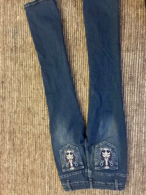 Blå jeans med broderade bakfickor - Säljer ett par blå jeans med raka ben och snygga broderade detaljer på bakfickorna i vitt och ljusblått. Klassisk femficksmodell med normal midja och kontrastsömmar. Perfekta för dig som vill ha något unikt i garderoben!