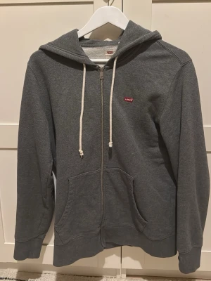 Grå zip hoodie från Levi's - Mörkgrå hoodie från Levi's med dragkedja, vita snören i huvan och klassisk röd Levi's-logga på bröstet. Mjuk bomullsblandning, kängurufickor framtill och ribbade muddar vid ärmar och nederkant. Perfekt för en avslappnad och stilren look. Är lite större i modellen. Passar någon som bär M