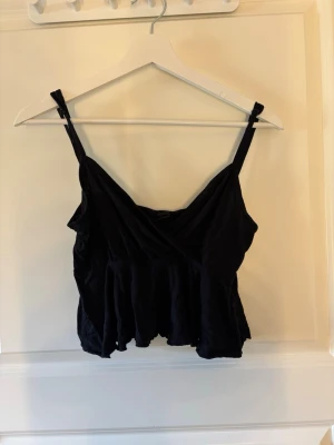 Svart omlott linnetopp - Snygg svart linnetopp med omlott-detalj och smala axelband. Toppen har en lätt volang nertill och är perfekt för en trendig och avslappnad look. Materialet känns mjukt och lätt mot huden.