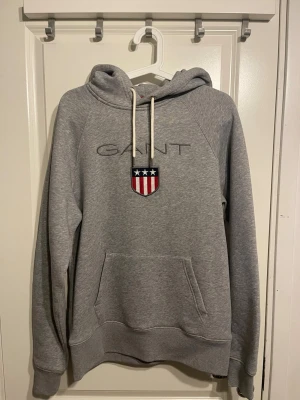 Grå hoodie från GANT med logga - Snygg grå hoodie från GANT med broderad logga och amerikanskinspirerat sköldmärke på bröstet. Klassisk känguruficka framtill, vita dragsnören i huvan och mjukt material. Perfekt för en avslappnad och stilren look.