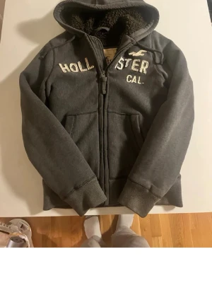 Hollister hoodie - Mysig mörkgrå hoodie från Hollister med broderad logga och text på bröstet. Dragkedja framtill, två fickor och fluffigt foder invändigt.  Strl Medium men skulle säga den sitter mer som en Large