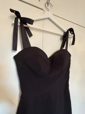 Svart klänning med knytband - En svart, figurnära klänning med knytband på axlarna och markerad byst. Klänningen har en enkel och stilren design med en feminin siluett och är perfekt för dig som gillar klassiska plagg med en twist. Materialet känns mjukt och följsamt. Köpt för 1000kr.